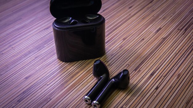 A pair of sleek, modern wireless headphones resting on a wooden desk next to a smartphone displaying a music streaming app. The headphones are black with silver accents, and the background is blurred to focus on the product.